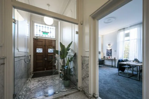 Entreehal met marmeren vloer en houten deur, met zicht op een modern ingericht kantoor aan de Van Eedenstraat.